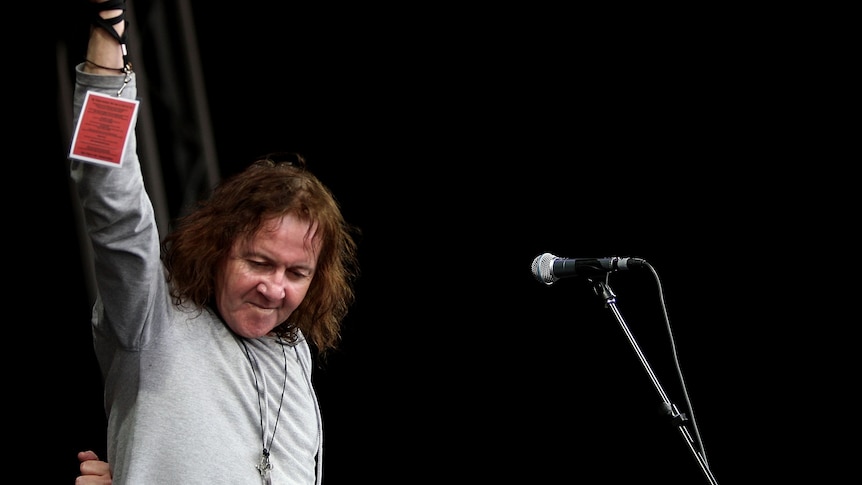 Chris Bailey of The Saints holds one arm aloft and looks down while performing on stage. He has long hair and wears a grey shirt