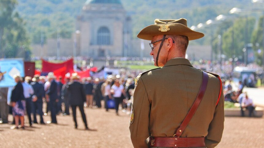 A man stands in uniform looking at the crowds gathered for the national Anzac Day march with the War Memorial in the distance.