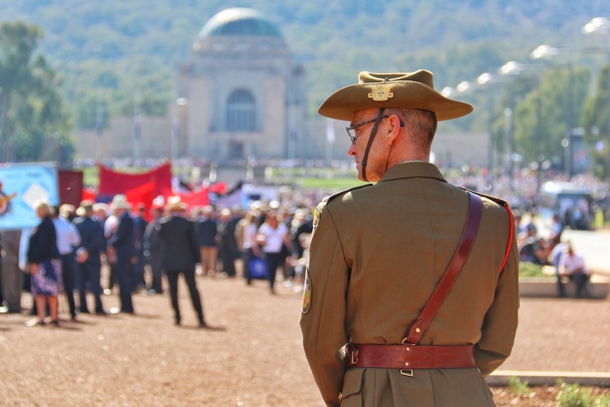 A man stands in uniform looking at the crowds gathered for the national Anzac Day march with the War Memorial in the distance.