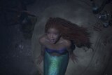 A black girl with red hair and a green tail dressed as Ariel the mermaid is looking up and singing with her mouth open. 