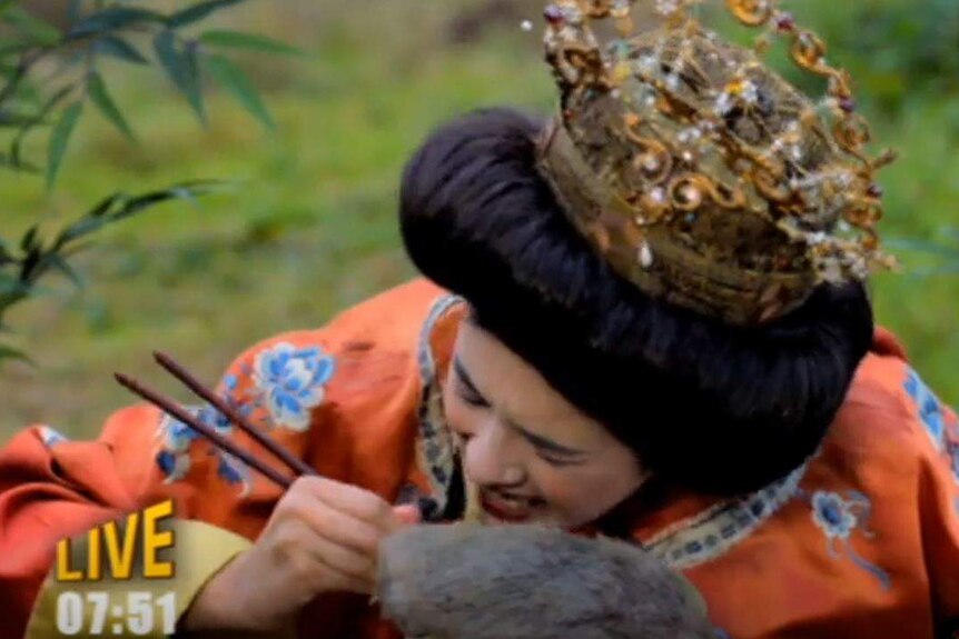 An actor made up to play an ancient Chinese empress pretends to use chopsticks to eat a rat while sitting among bamboo.