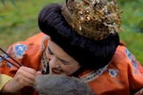 An actor made up to play an ancient Chinese empress pretends to use chopsticks to eat a rat while sitting among bamboo.