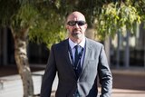A man with a shaved head and neat beard, wearing dark sunglasses and a suit, walks through a court complex.