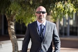 A man with a shaved head and neat beard, wearing dark sunglasses and a suit, walks through a court complex.
