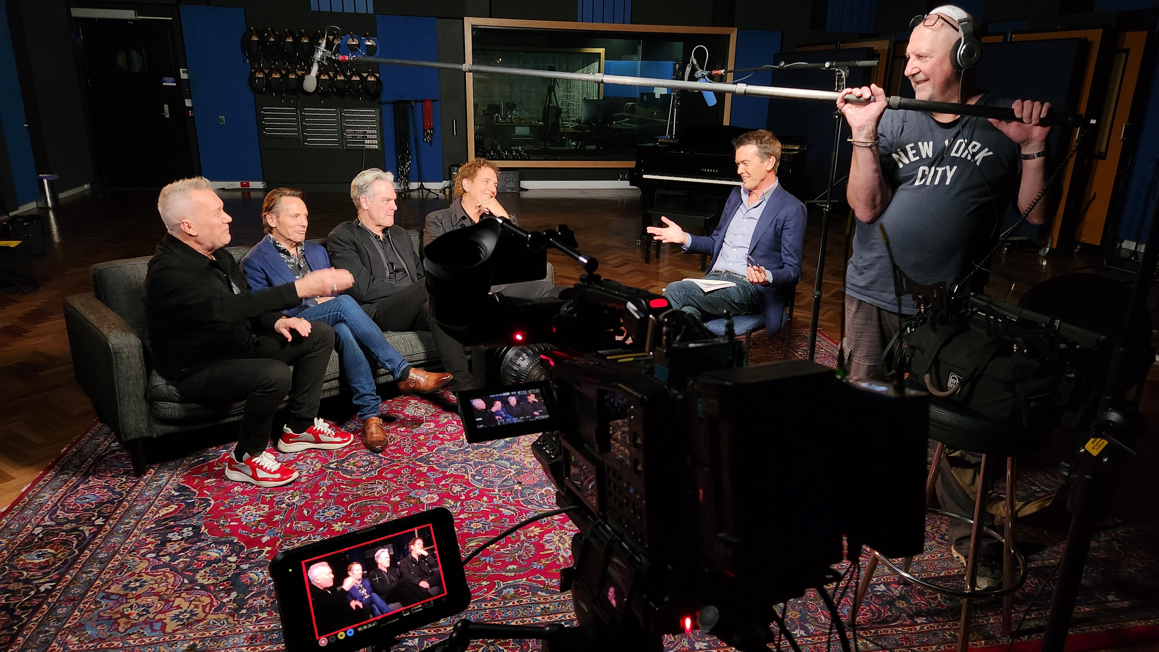 TV interview scene. Band of four men left of frame, journalist and sound op to the right. Camera out of focus in centre.