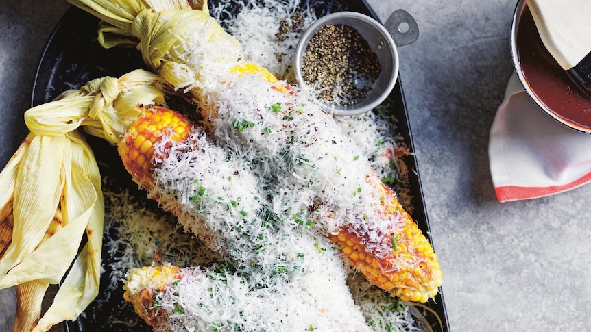 Three corn cobs with silks attached covered in snowy cheese on a grill pan set on a table.