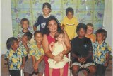 A woman with long dark hair sits surrounded by a dozen young, smiling children