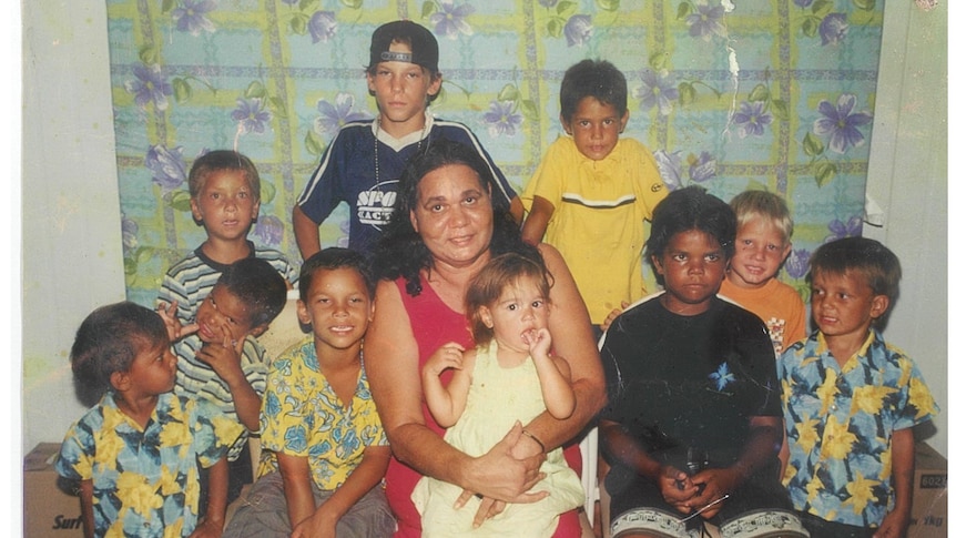 A woman with long dark hair sits surrounded by a dozen young, smiling children
