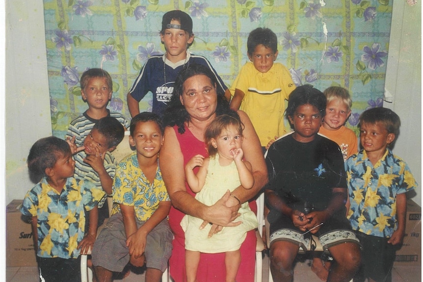 A woman with long dark hair sits surrounded by a dozen young, smiling children