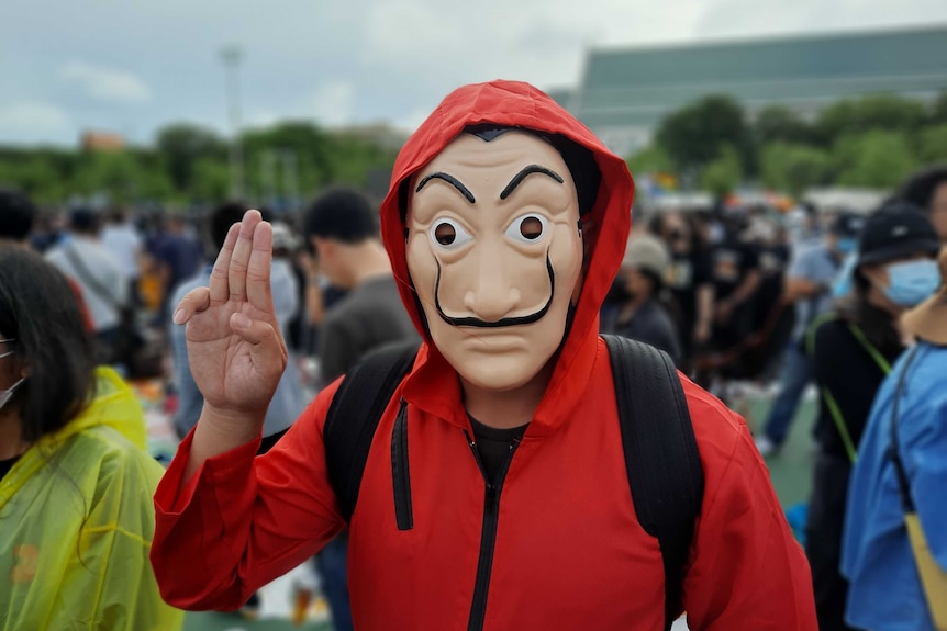 A person in a red hoodie and anonymous mask doing a three-finger salute.