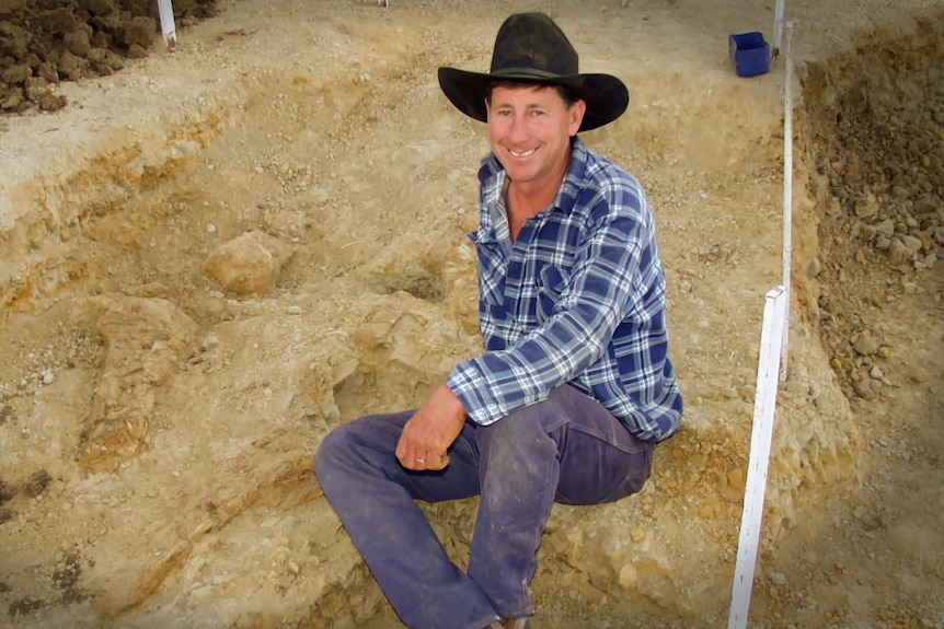 A man sits on the ground with a dug up patch of dirt and dinosaur bones.