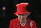 Queen Elizabeth wearing a red suit standing next to a priest by a car.