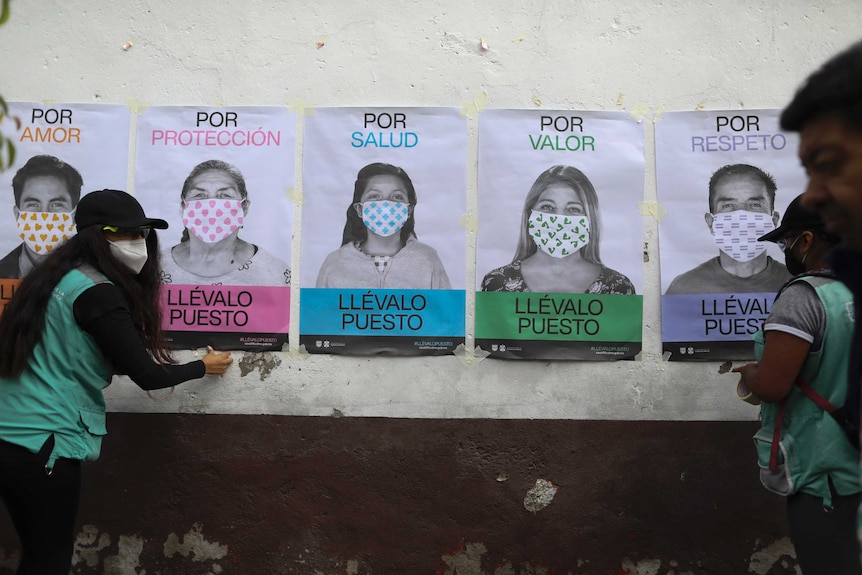 People wearing face masks fix posters bearing slogans in Spanish encouraging people to wear face masks to a white wall