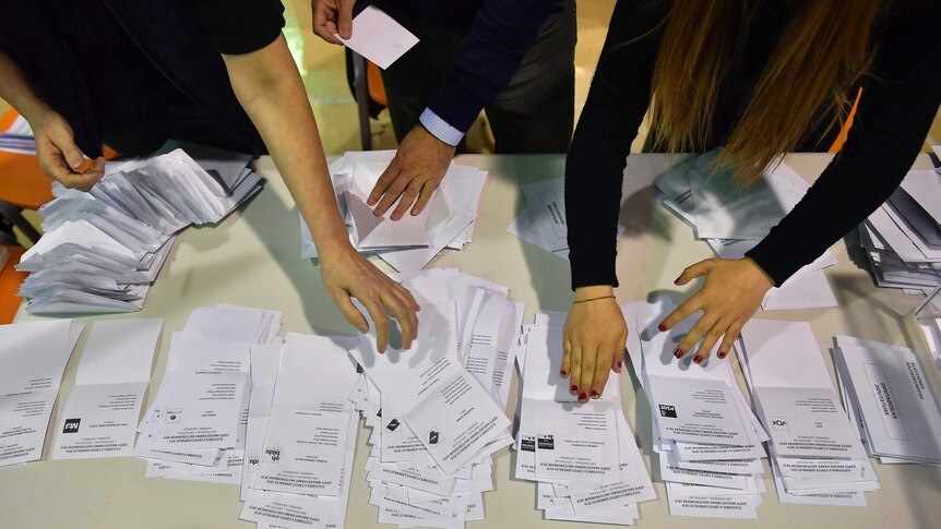White ballot papers sorted into piles by three people, only their hands are visible.
