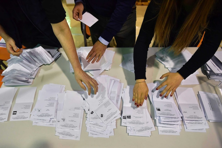 White ballot papers sorted into piles by three people, only their hands are visible.