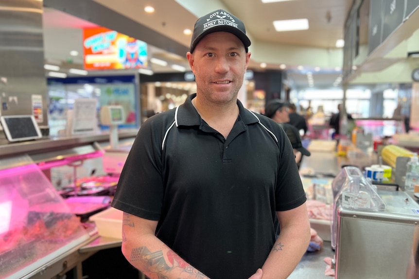 A tattooed man wearing a black shirt and  badged cap stands inside a butcher's shop
