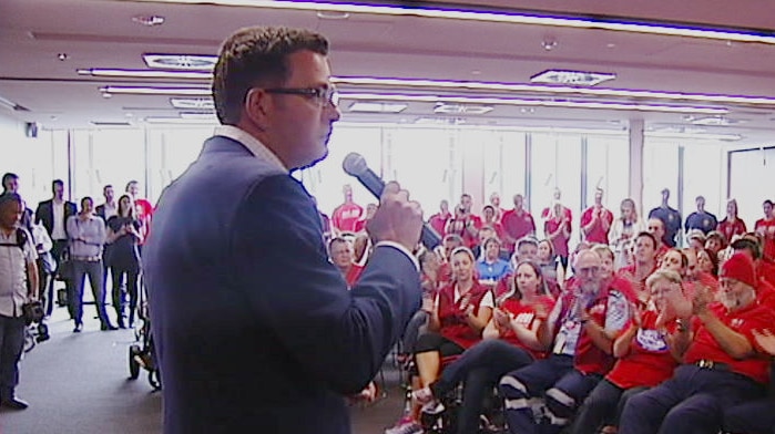 Daniel Andrews speaks into a microphone in a roomful of people in red shirts.