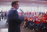 Daniel Andrews speaks into a microphone in a roomful of people in red shirts.