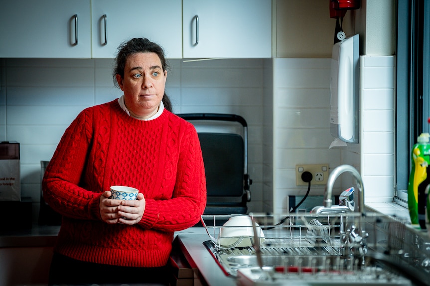 Fiona McKenzie stands in her kitchen and looks out the window while holding a cup in both hands.