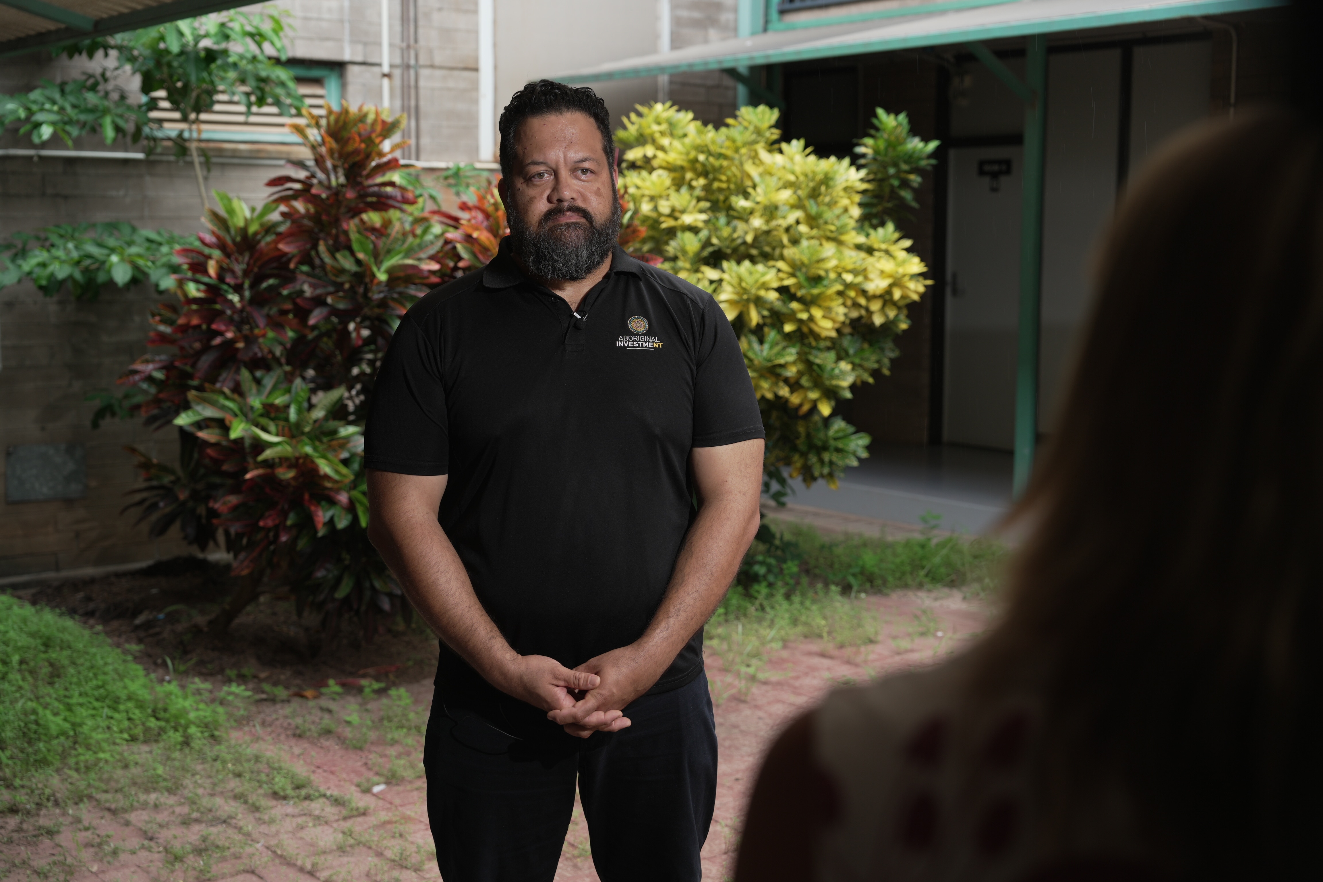 A man in black polo shirt stands in a courtyard and speaks to a reporter.