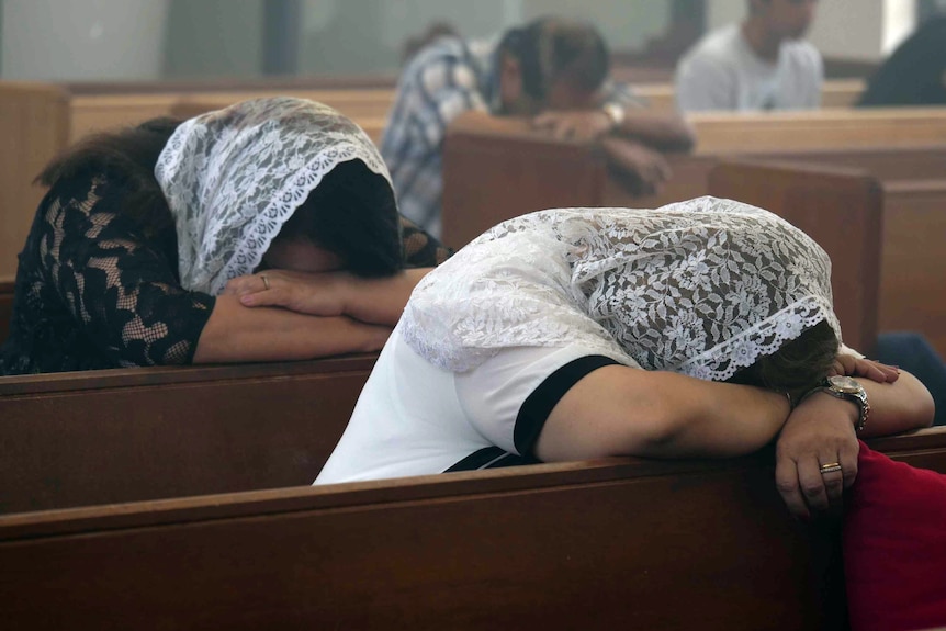 Two women with lace scarfs over their heads sit in a church pew, with their heads resting on their arms in prayer.