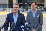 Two men wearing suits stand in front of microphones with Adelaide Oval behind them