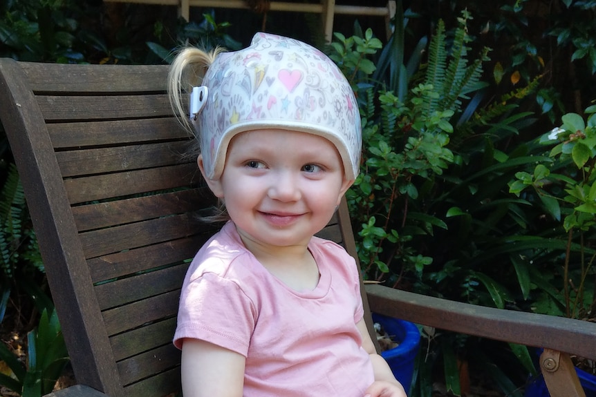 A toddler in a pink shirt and white helmet sits outside