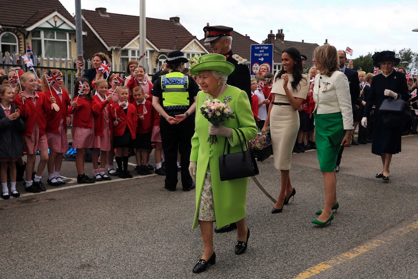 Queen Elizabeth II and Meghan, the Duchess of Sussex, walk along a street with kids waving.