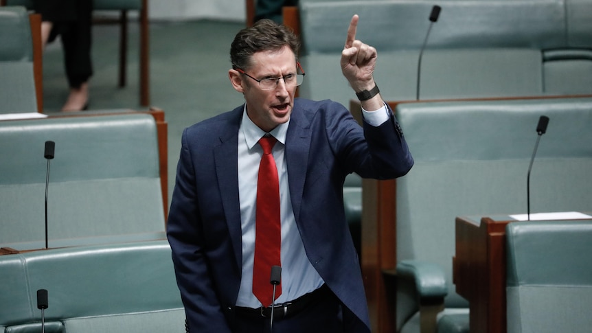 Jones is standing wearing a blue suit and red tie, left finger in the air.