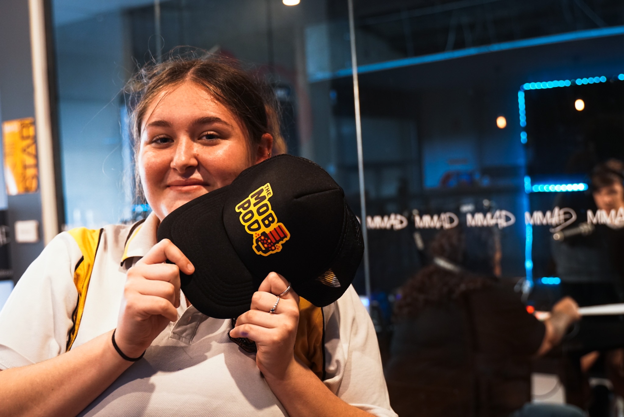 A young woman holding a cap with the words "Mob Pod" and smiling, two people working inside a booth can be seen.