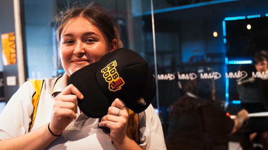 A young woman holding a cap with the words "Mob Pod" and smiling, two people working inside a booth can be seen.