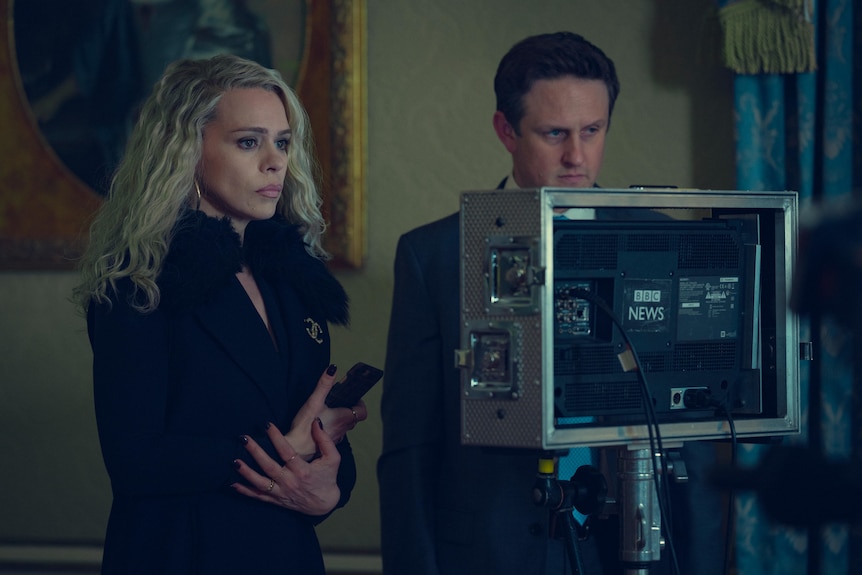 A film still of Billie Piper, clutching a mobile phone, standing at a TV monitor with Richard Goulding.