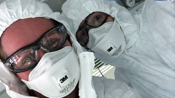 Two people wear personal protective equipment including masks, goggles, gloves and suits.