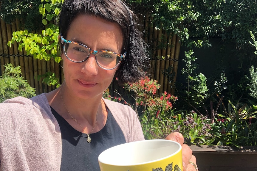 A woman looks into the camera holding a tea cup in front of her. 