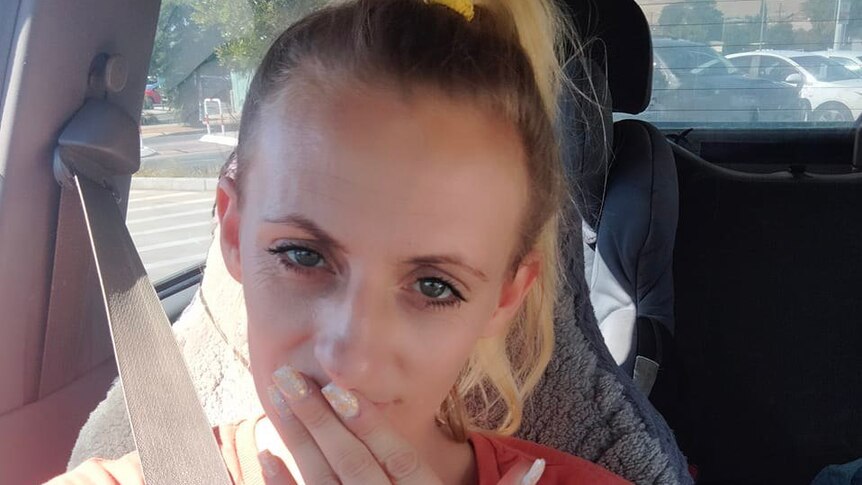 A woman with a blond ponytail in an orange top with her fingers touching her mouth