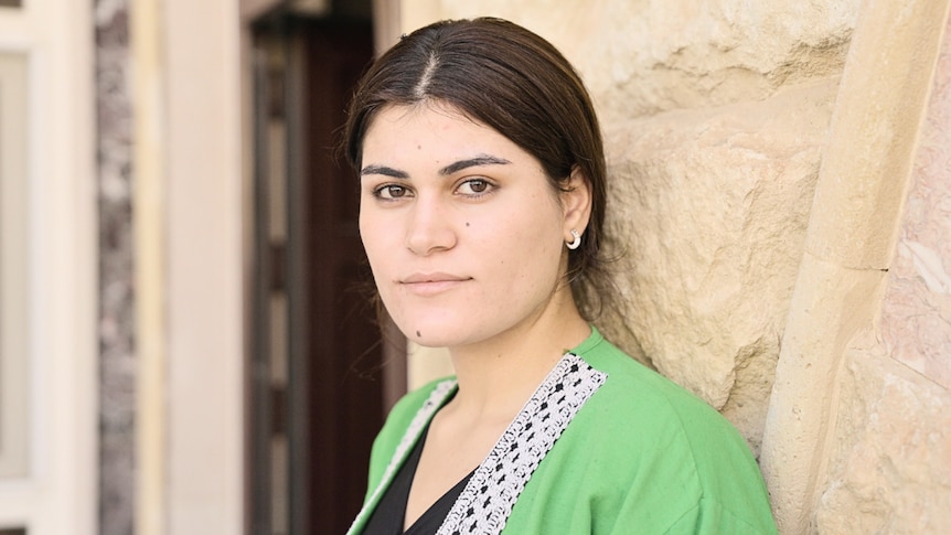 A woman in a green top stands against a stone wall.