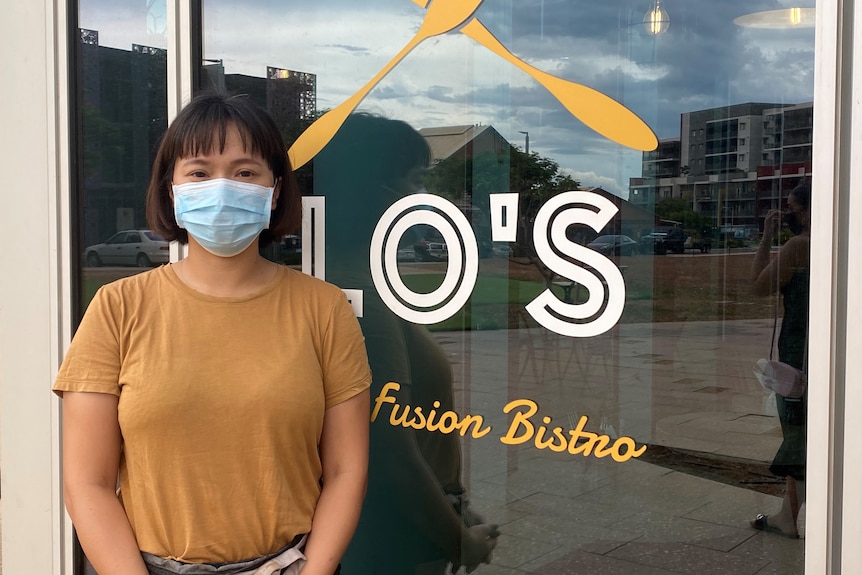 A woman wearing a blue mask and a yellow shirt stands in front of a window with 'Lo's' written on it