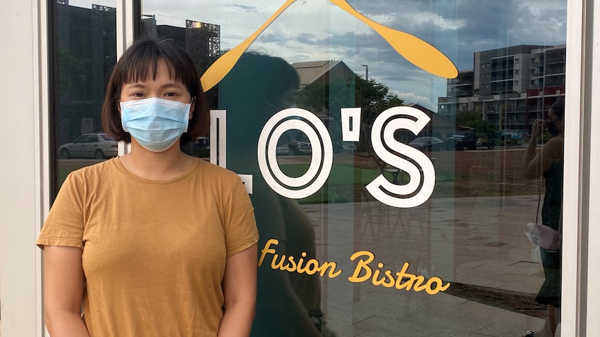 A woman wearing a blue mask and a yellow shirt stands in front of a window with 'Lo's' written on it