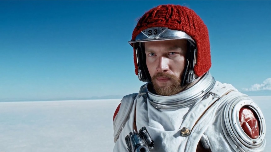 A realistic-looking man stares solemnly into the distance, standing in a salt desert wearing a space suit.