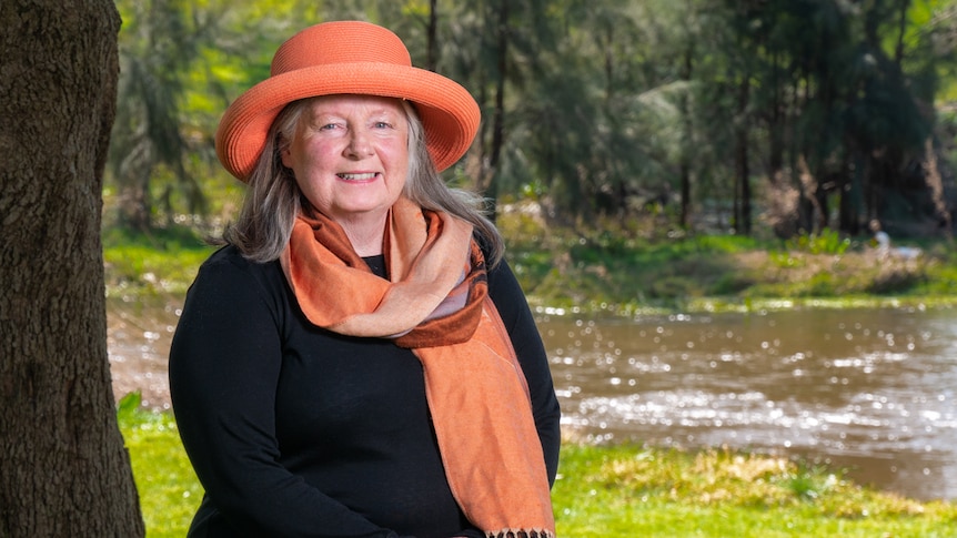 An older woman wearing a peach-coloured hat and matching scarf stands in front of a river.