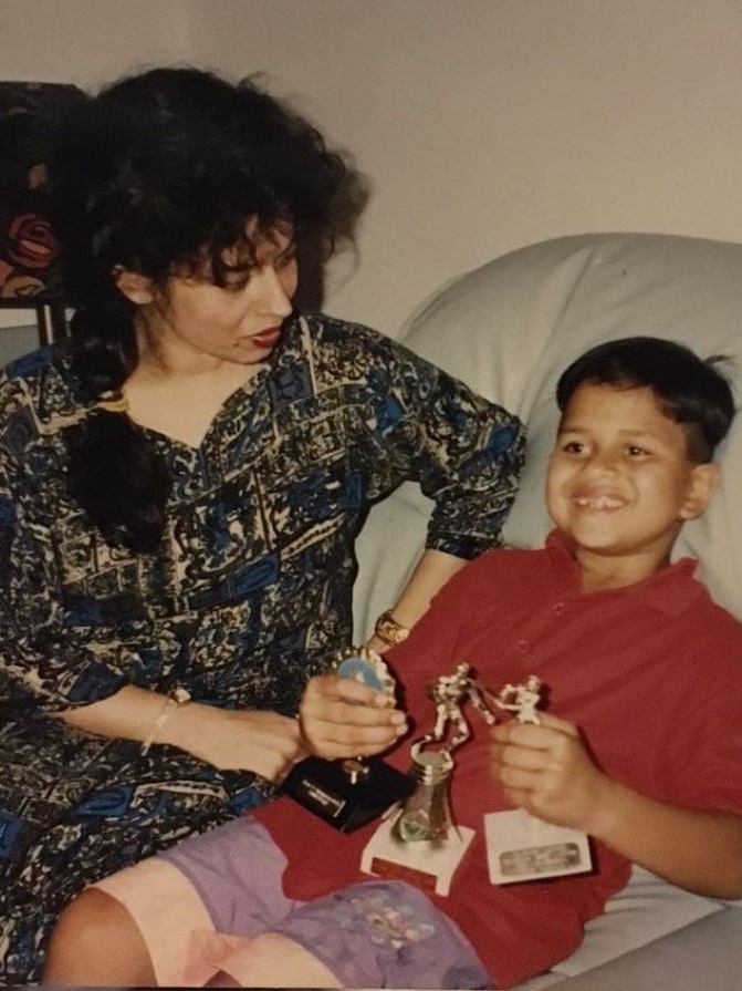 A young Usman Khawaja holding three sporting trophies while sitting next to his mother