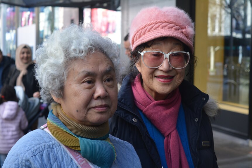 Two elderly women of Asian descent look are pictured close-up while one wears a pink furry hat.