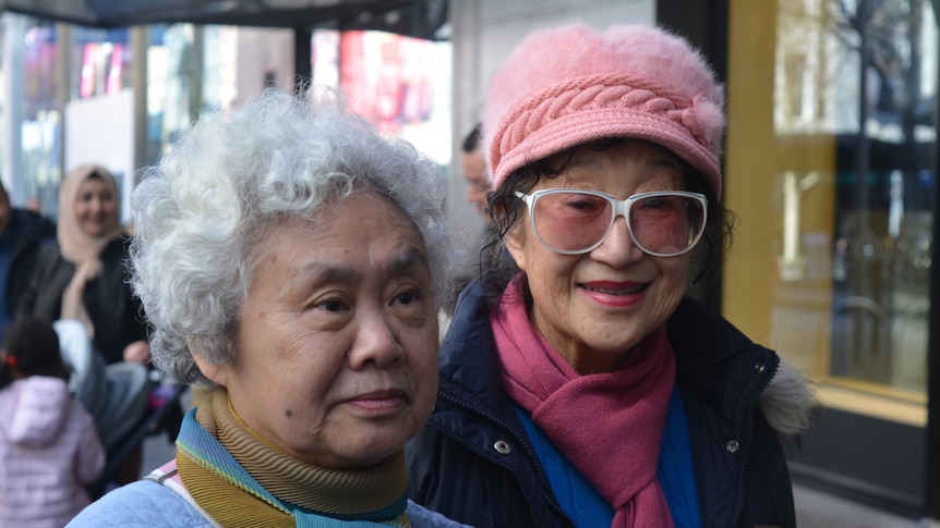 Two elderly women of Asian descent look are pictured close-up while one wears a pink furry hat.