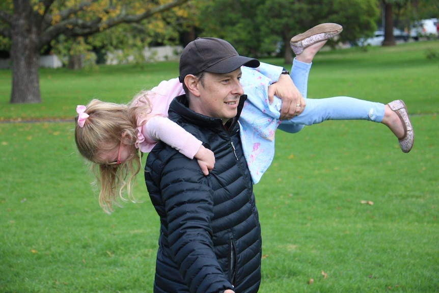 A dad wearing a cap and black jacket in a park holds his daughter over his shoulder.