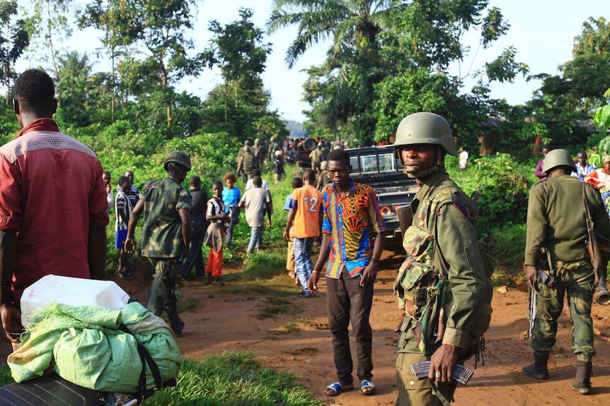 Against a lush green landscape, DRC Congo soldiers patrol a dirt road with a man in a bright shirt staring at the camera.