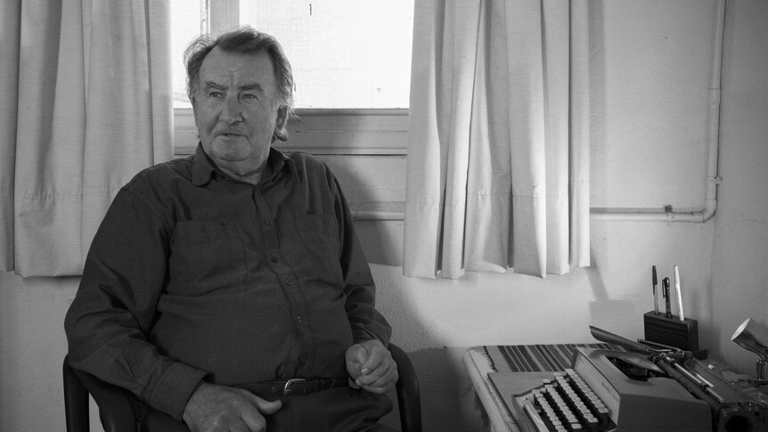Gerald Murnane sitting in front of a window, at a desk with a typewriter and pens.