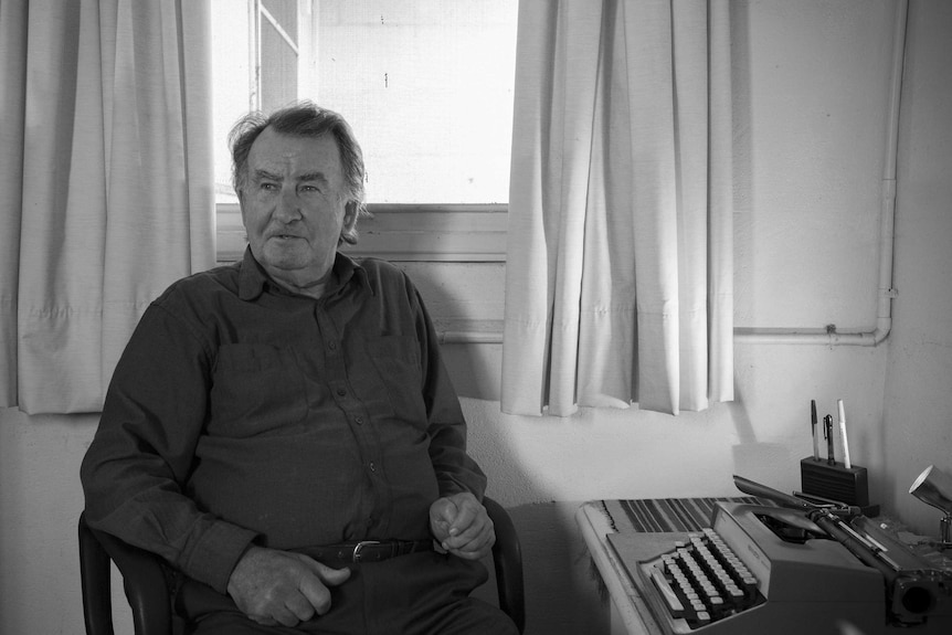 Gerald Murnane sitting in front of a window, at a desk with a typewriter and pens.