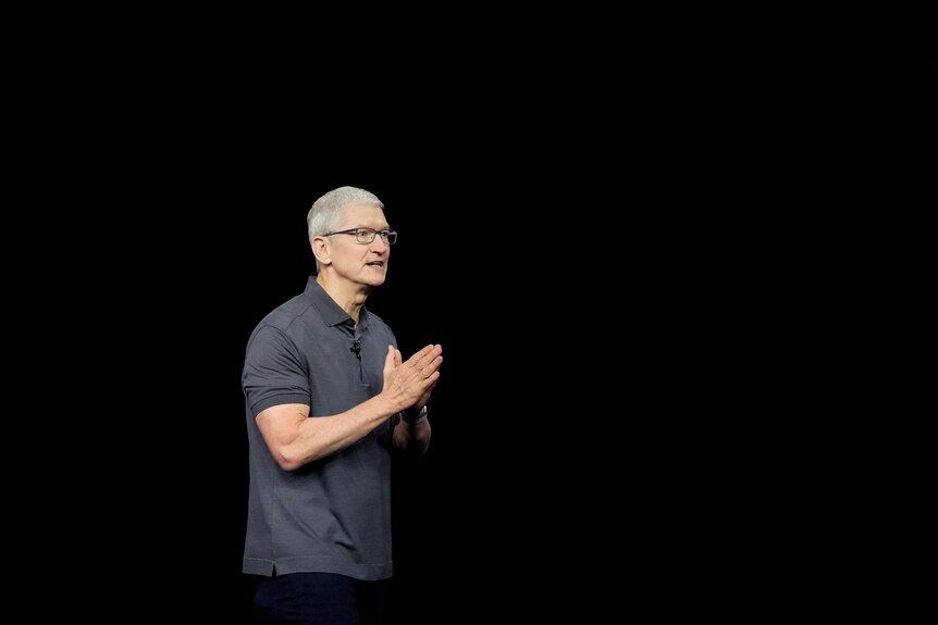 Tim Cook stands on a stage wearing a polo t-shirt and glasses, speaking and holding his hands together