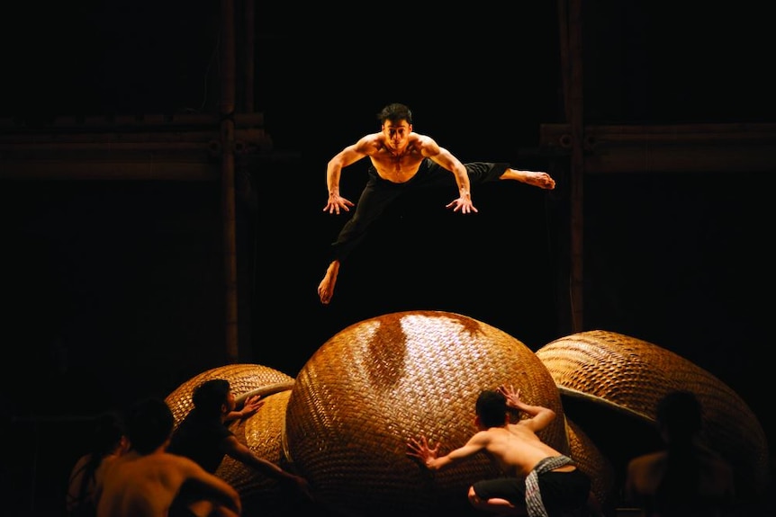 A male acrobat leaps over a huge woven orb.