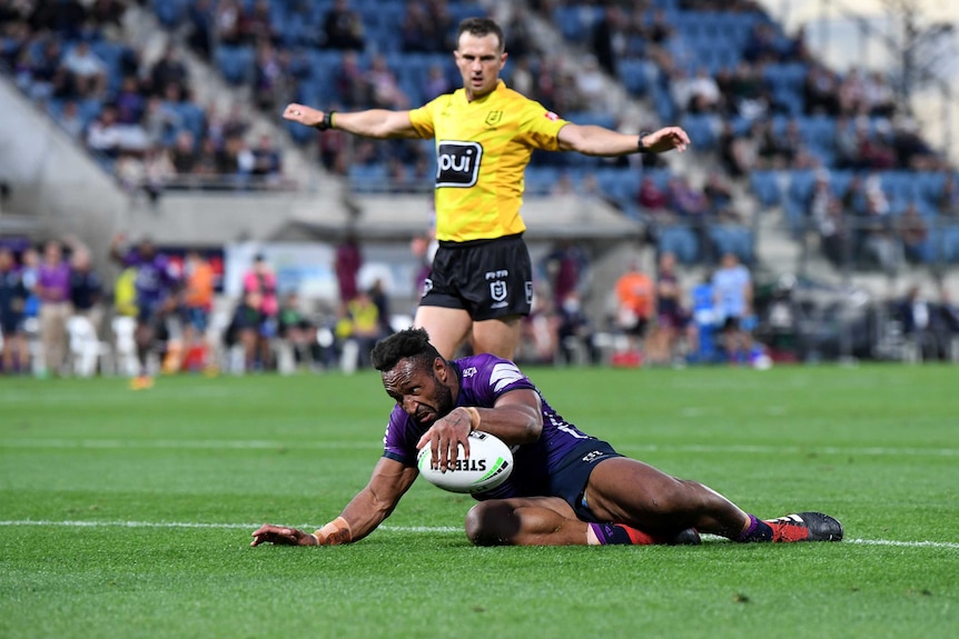 A Melbourne Storm NRL player looks to ground the ball for a try while on his knees as the referee stands in the background.
